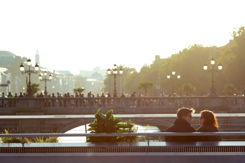 young-couple-sitting-on-bench-with-bridge-in-background